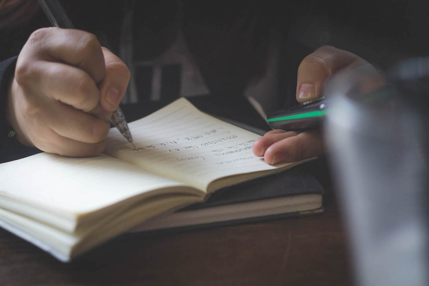 A man writing in a notebook while looking at his phone